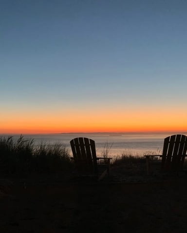 Two Adirondack chairs silhouetted against a vivid orange and blue sunset over the Puget Sound shoreline